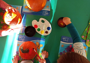 Na stoliku leżą dynie oraz paleta z farbami, dzieci kolorują farbami swoje warzywo.